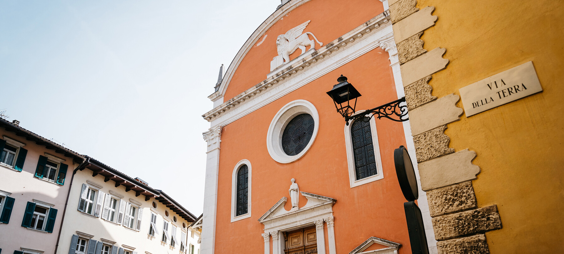 Façade of the Church of San Marco, in the historic center of Rovereto. The protagonists of the image are the colors: the orange of the church, the yellow ochre of the building in the foreground, on which “Via della Terra” is written, and the white and soft pink of the buildings in the background. Although not inhabited by people, it is a lively, joyful image.