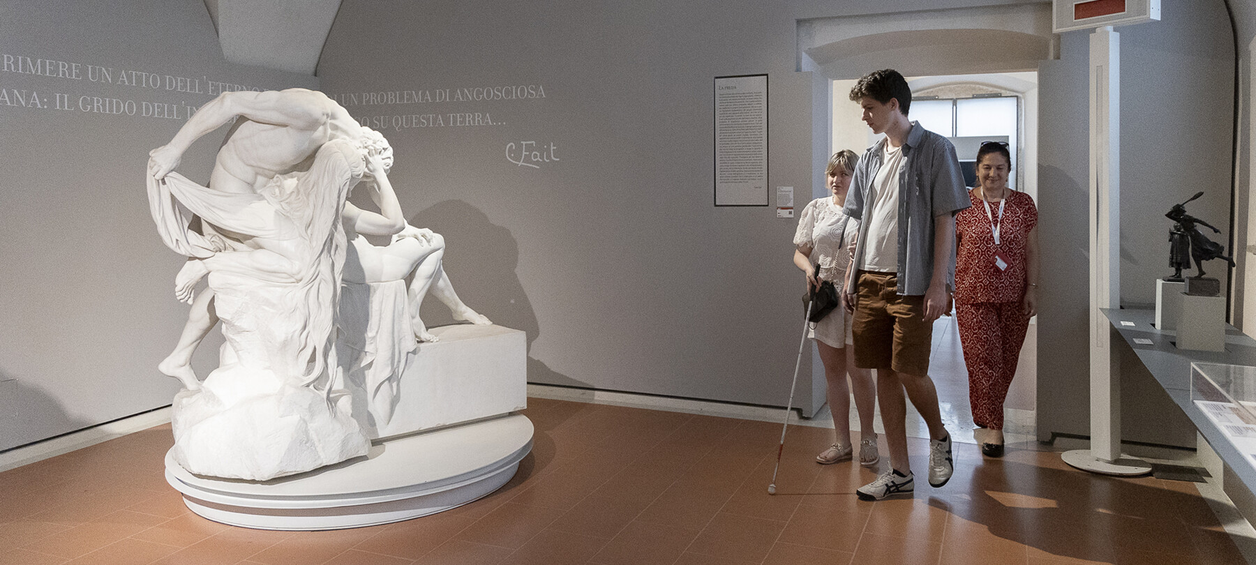 Carlo Fait Hall of the Palazzo Alberti Poja, in Rovereto. On the left is a plaster sculpture depicting a man assaulting and oppressing a woman. Three people enter through the door on the right: a boy, a blind woman, and the tour guide accompanying them. 