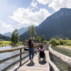 A woman in a wheelchair and a tour guide walk along a catwalk suspended over water. Their backs are turned and they seem to be chatting with each other. It is summer, a beautiful sunny day. The green mountains are reflected on the surface of the water. 