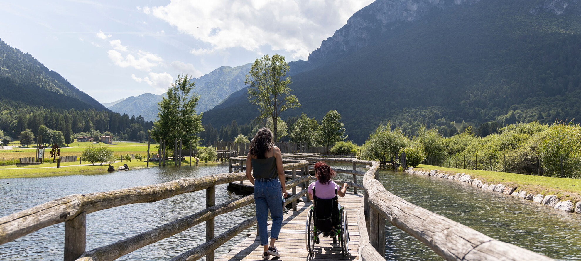 A woman in a wheelchair and a tour guide walk along a catwalk suspended over water. Their backs are turned and they seem to be chatting with each other. It is summer, a beautiful sunny day. The green mountains are reflected on the surface of the water. 