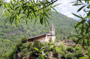 Church of Santo Stefano di Carisolo. The church stands atop a granite cliff. Behind, the forest climbs up the side of the mountain: the green of its trees mingles with that of the leaves that, blurred in the foreground, frame the image. On the south-facing side of the church, the one immortalised in the photograph, frescoes can be recognised in the distance. The church has a small stone bell tower and, to its right, three wooden crosses stand out.  