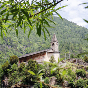 Church of Santo Stefano di Carisolo. The church stands atop a granite cliff. Behind, the forest climbs up the side of the mountain: the green of its trees mingles with that of the leaves that, blurred in the foreground, frame the image. On the south-facing side of the church, the one immortalised in the photograph, frescoes can be recognised in the distance. The church has a small stone bell tower and, to its right, three wooden crosses stand out.  