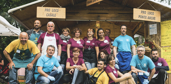 Gruppenfoto mit einigen Freiwilligen der Veranstaltung Tutti #fuori (alle #nachdraußen), dem großen Wanderfestival der Pro Loco im Trentino. Die Farben der T-Shirts, die sie tragen, verraten uns, dass es sich um Freiwillige von verschiedenen Pro Locos handelt. Aber hier arbeiten sie zusammen, und gemeinsam feiern sie ihr Engagement.