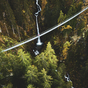 Ponte sospeso in Val di Rabbi. Nell’immagine, il lungo ponte tibetano è ripreso dall’alto. Sotto svettano i larici e sullo sfondo una cascata si tuffa tra le rocce e gli alberi. Una persona sta attraversando il ponte, la si nota per la sua giacca rossa che spicca tra i colori più tenui della natura che la circondano.