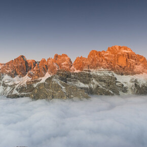 The picture is taken from the summit of Paganella: one can see the snow-capped Brenta Dolomites at dawn coloured pink by the phenomenon of alpenglow; below, clouds frame the picture.