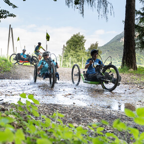 Siamo sull’Altopiano della Vigolana. È una bella giornata estiva, luminosa. Quattro turisti, amanti dell’attività outdoor, fanno biking seguendo un sentiero sterrato: tre di loro guidano una handbike. Insieme, si godono il sole sulla pelle, l’aria fresca e il verde dei boschi che li circonda. | © ©Trentino Marketing_Gianluca Prati_Accessibilita_Vigolana_2024_00012